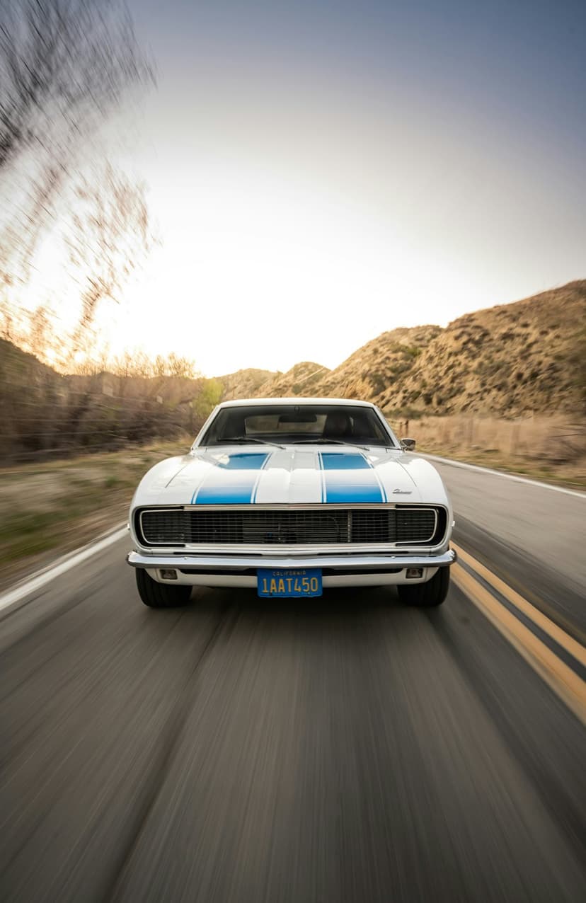 White Chevrolet Camaro with blue stripes racing on a highway at sunset with mountains in the background.