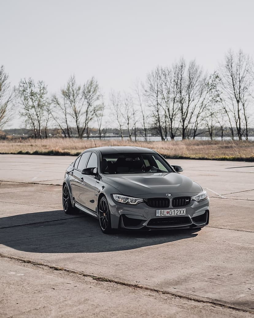BMW M3 sedan in dark gray with black wheels parked on sunny asphalt near bare trees and a calm lake.