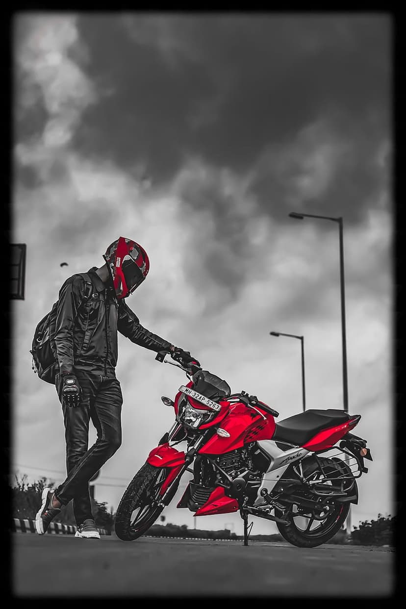 Man in a red helmet and black leather gear leans on a red motorcycle against a gray cloudy sky and streetlights.
