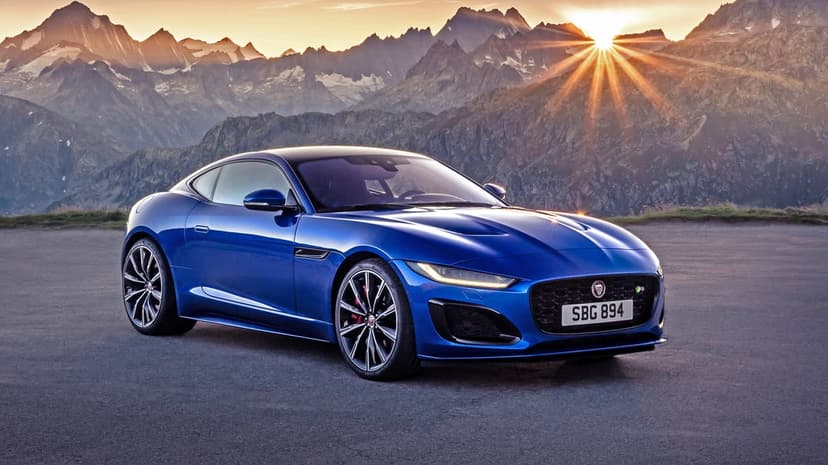 Blue Jaguar F-Type R coupe parked on asphalt with sun rays over snow-capped mountains at sunset.