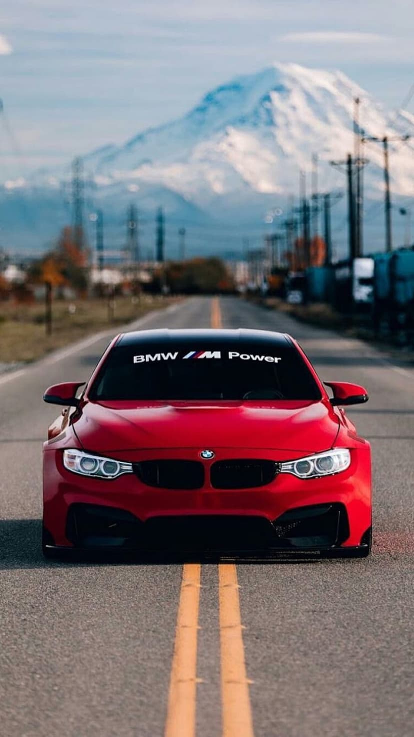 Red BMW M series sedan driving on a mountain road with snow-covered peaks and utility poles under clouds.