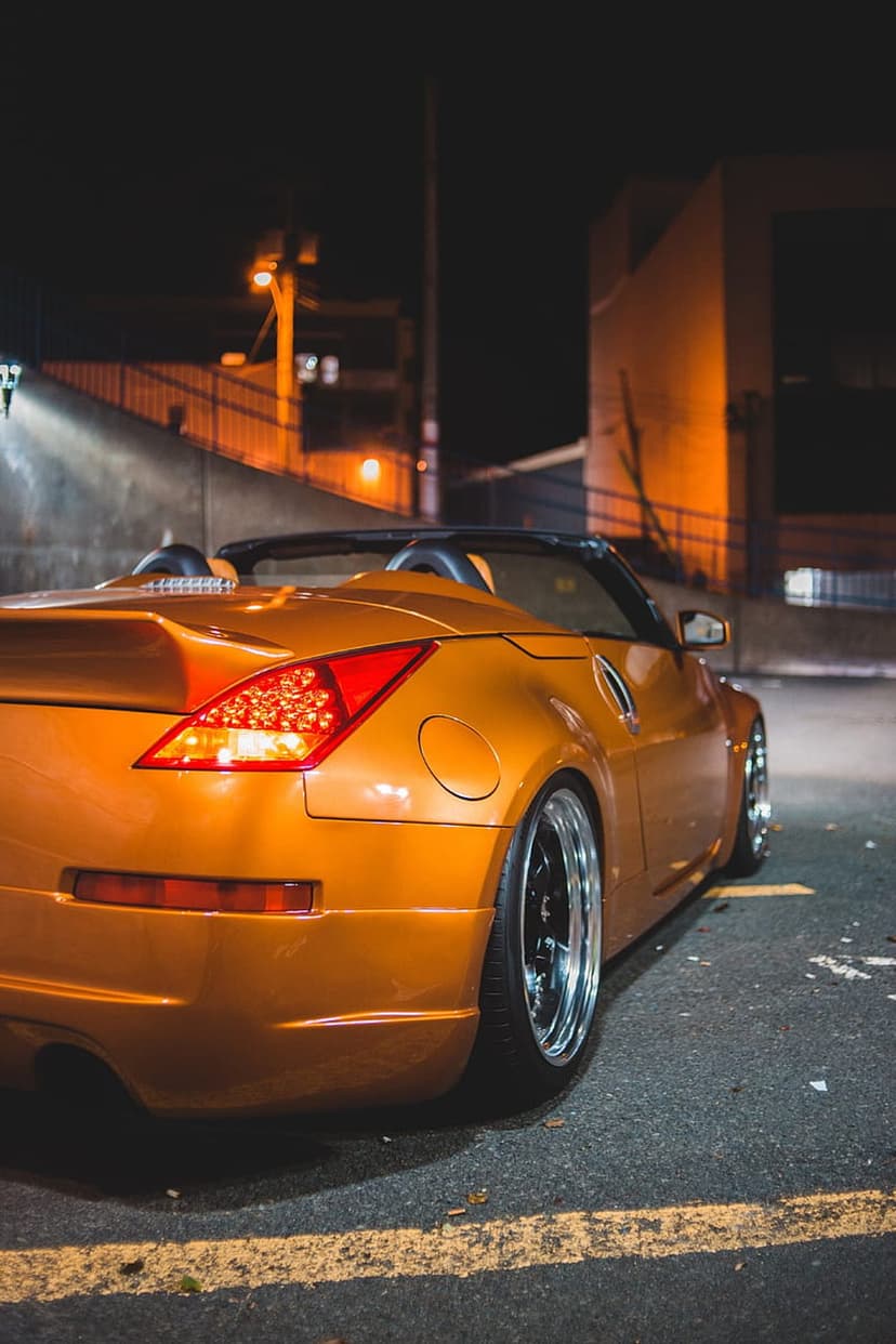 Orange convertible sports car with glowing taillights parked on a dark urban street under amber streetlights.