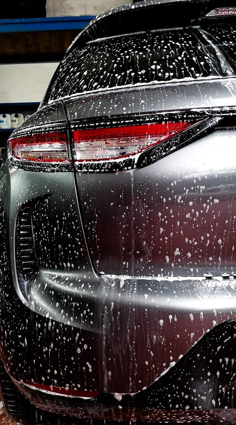 White soap suds drip over a gray car's rear taillight and metallic bumper during a detailed wash.