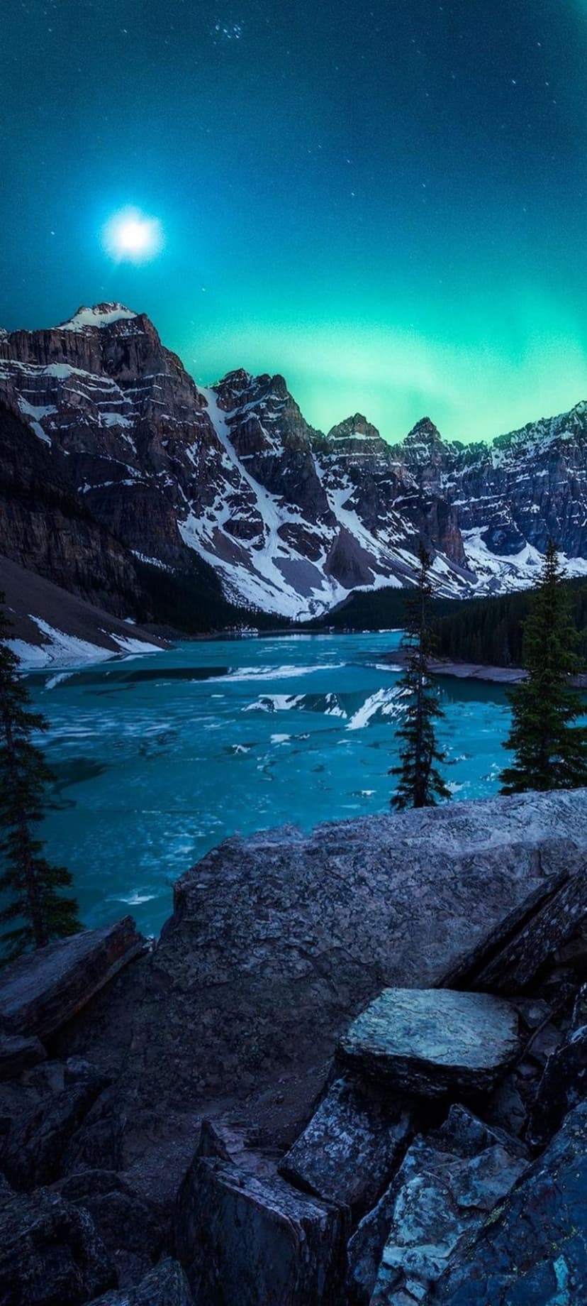 Aurora Borealis ribbons glow green over snow-dusted mountains and a still turquoise lake under a starry night sky.