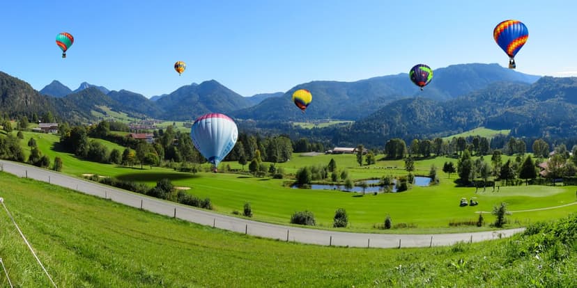 Multicolored hot air balloons soar above rolling green hills and winding roads under a clear, bright blue sky.