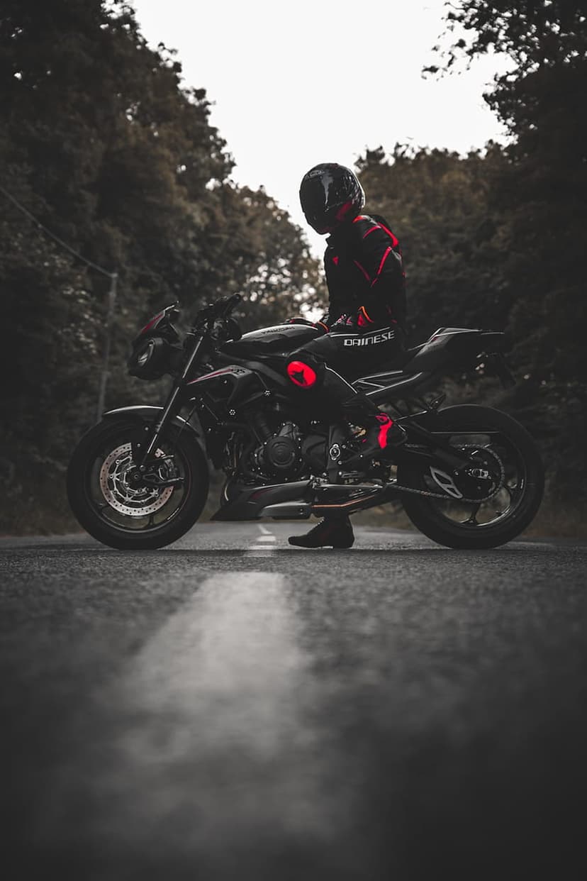 Motorcyclist in a Dainese suit sits on a black sportbike with red accents on a forest road under overcast skies.