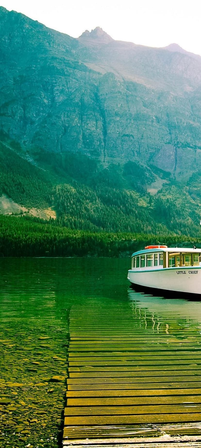 White boat named Little Chief cruises across a green lake toward tall mountains beside a rustic wooden dock.