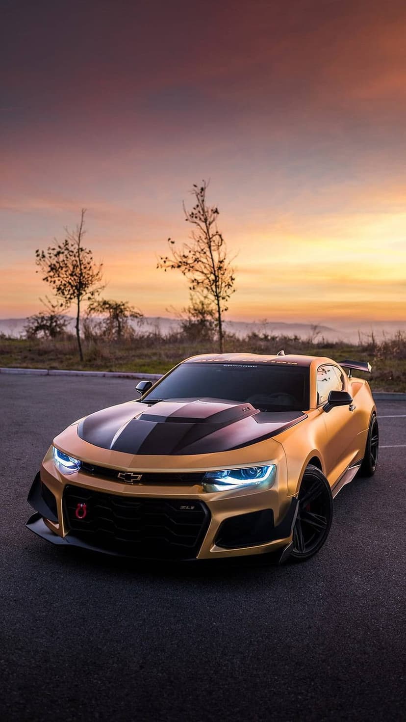 Golden Chevrolet Camaro ZL1 with racing stripes parked on asphalt against a vibrant purple and orange sunrise.