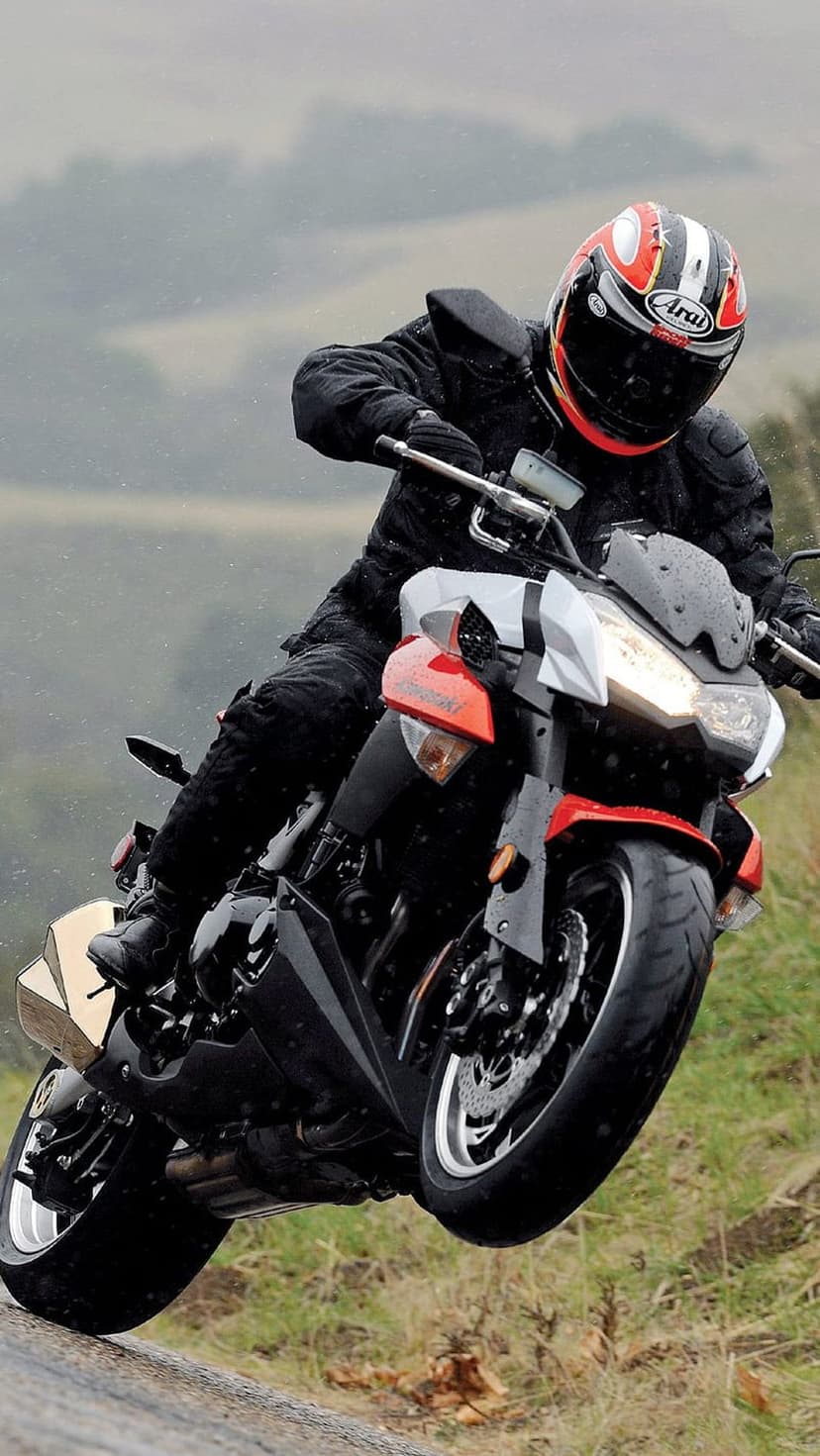 Motorcyclist in a red and black helmet leans a sportbike on a wet, rainy road with visible water droplets.