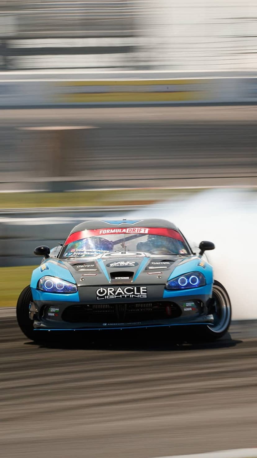 Blue Dodge Viper drifting on a race track with thick white tire smoke and glowing LED headlights in vertical view.