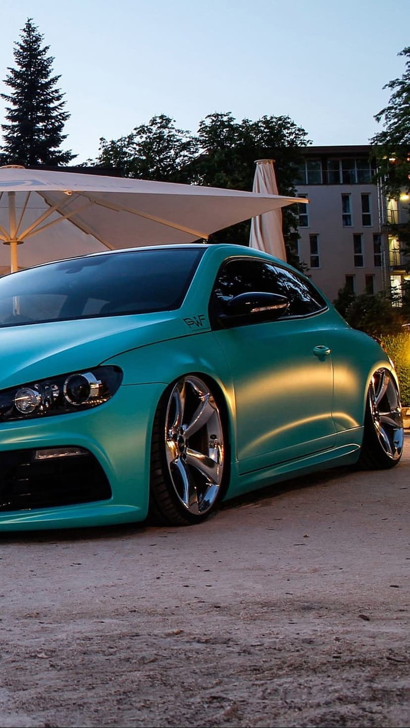 Mint green Volkswagen Scirocco with a slammed stance and chrome wheels parked in a modern urban outdoor setting.