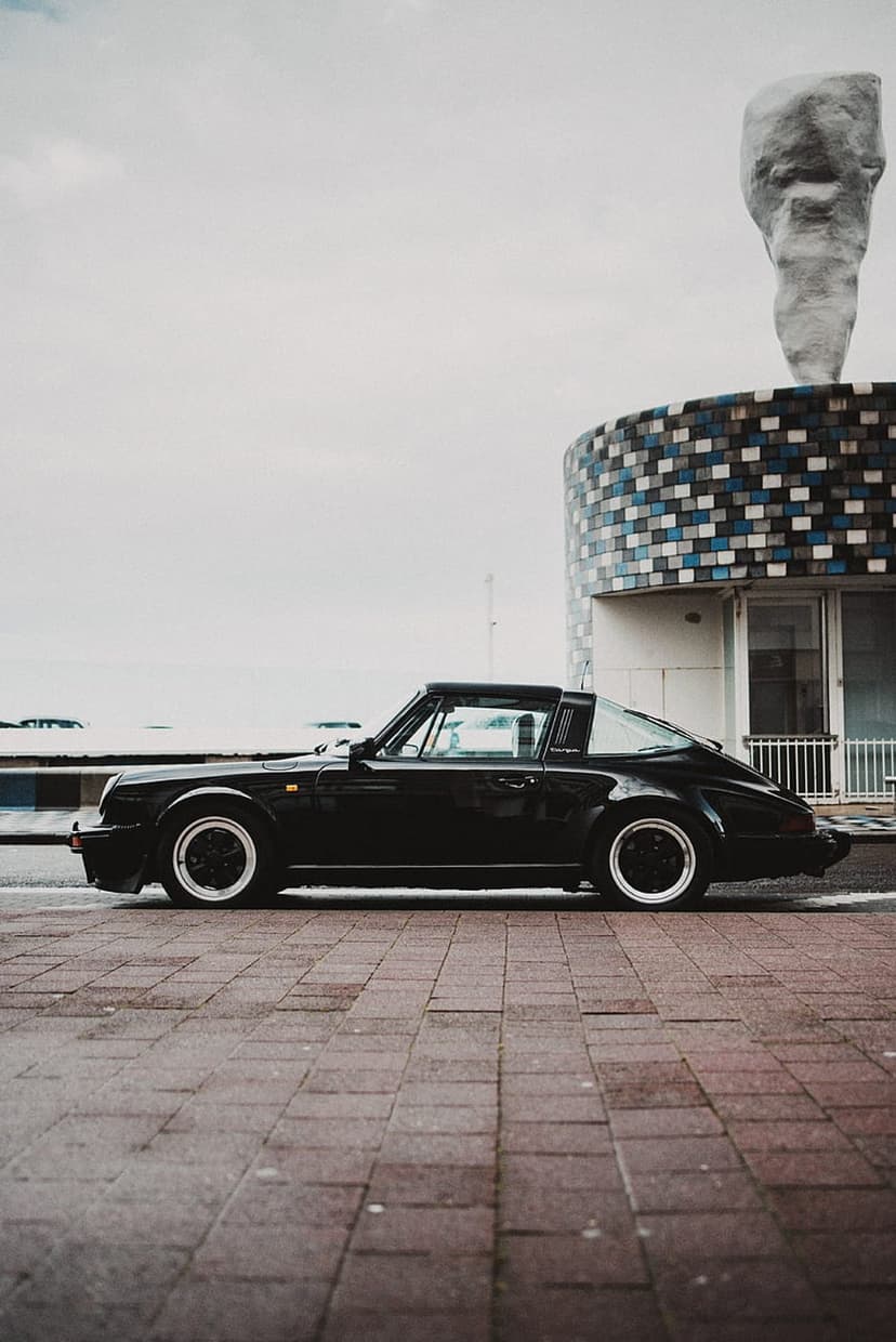 Black Porsche Targa on brick pavement near a blue mosaic building and abstract sculpture under gray skies.