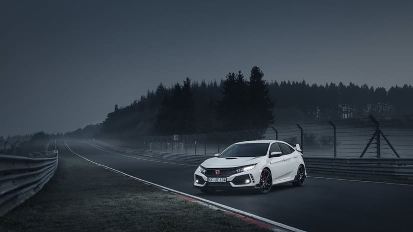 White Honda Civic Type R on a foggy race track at dawn with headlights on and a dark forest background.