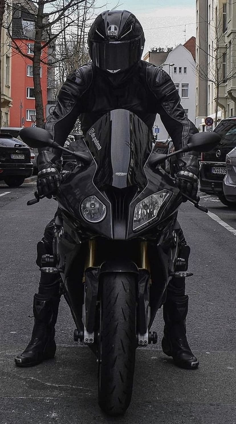 Motorcyclist in black leather gear sits on a sports bike with headlights on against a city street backdrop.