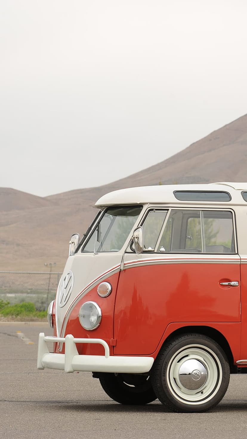 Red and white vintage VW bus front view with chrome bumper on an asphalt road with dry hills in the background.