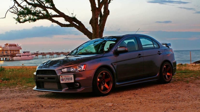 Grey Mitsubishi Lancer Evolution X with bronze wheels parked by a pier at sunset under a large tree.