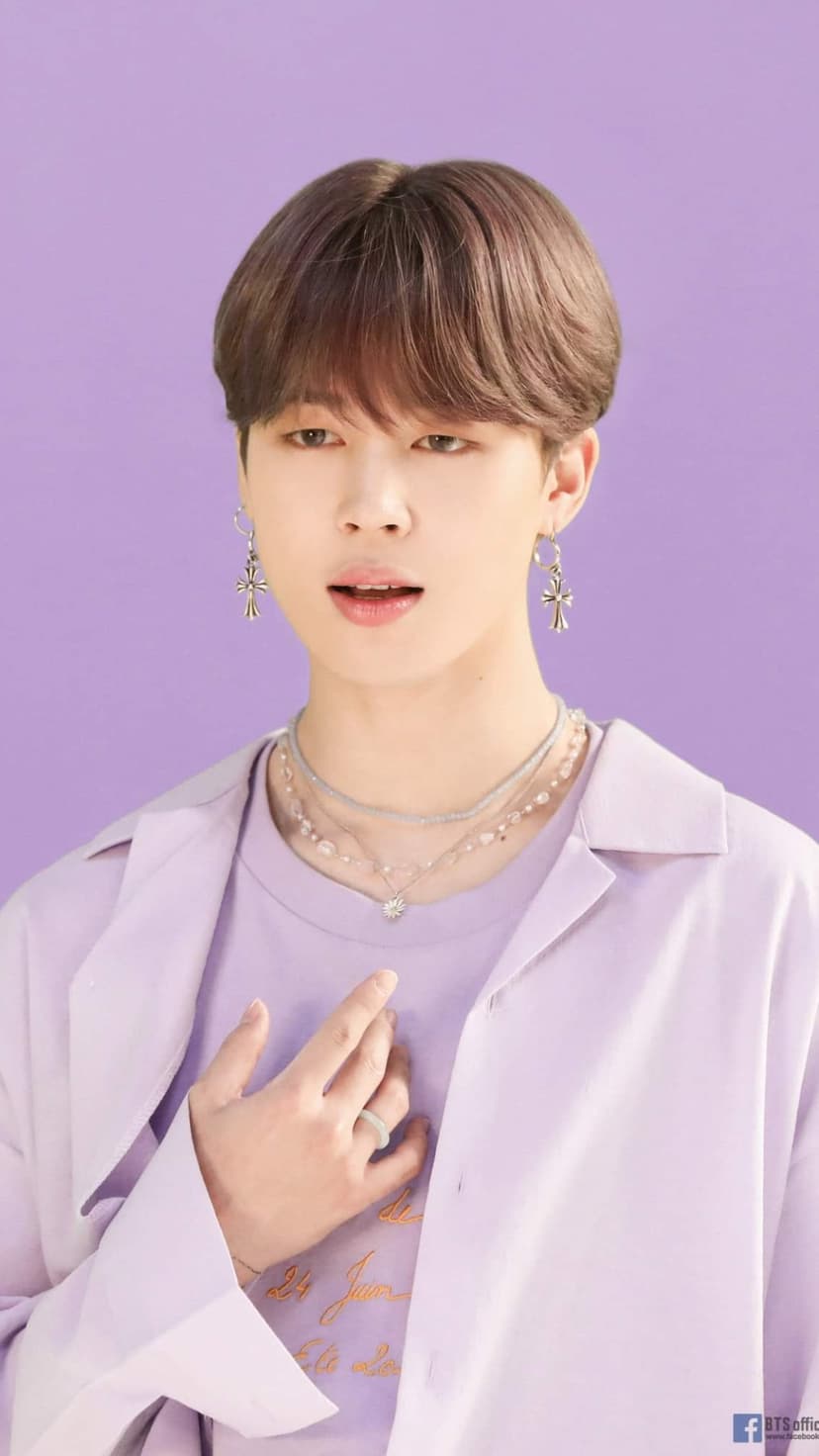 Park Jimin wears a purple shirt and cross earrings while resting his hand on his chest against a lavender backdrop.