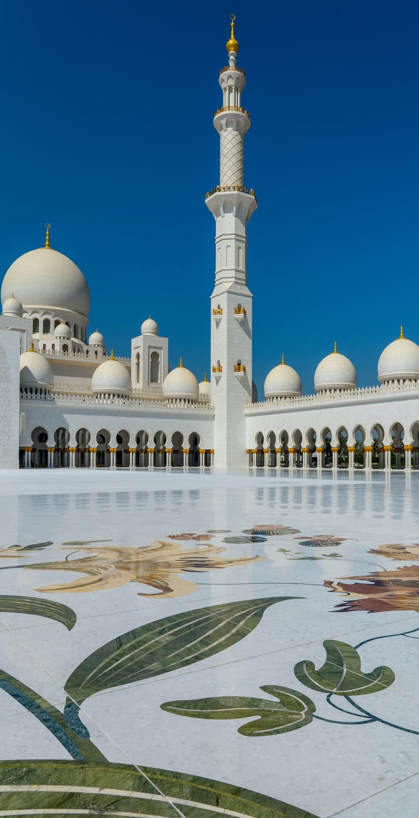 White marble courtyard with colorful floral stone inlays under a clear blue sky at the Grand Mosque in Abu Dhabi.