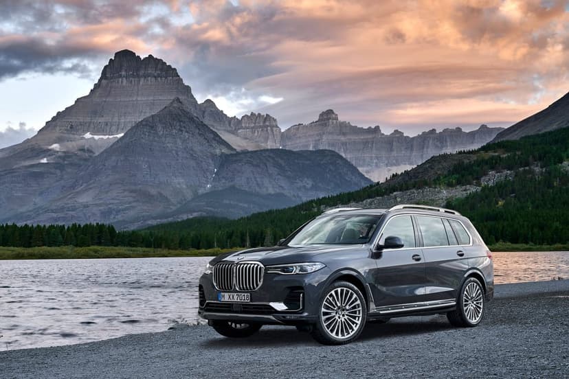 Dark grey BMW X7 SUV parked on a rocky lakeshore with jagged mountains and a sunset sky in the background.