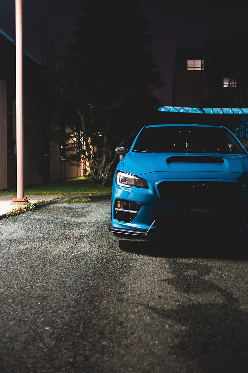 Blue Subaru WRX with an aggressive front splitter parked on asphalt under moody urban night lighting.