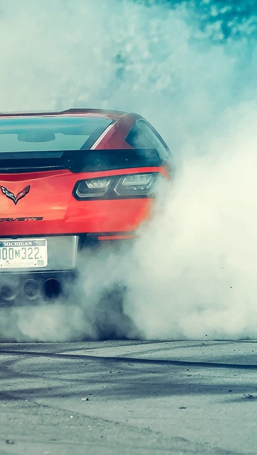 Red Corvette drifting on asphalt surrounded by white smoke with visible taillights and tire tread marks.