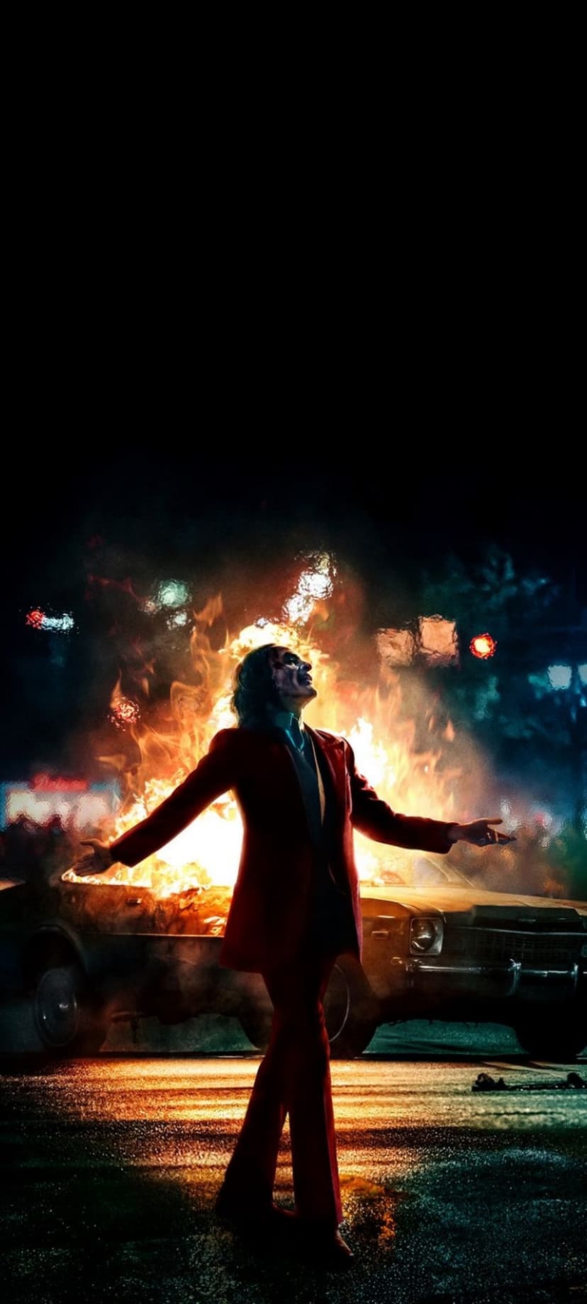 Joker in a red suit dances with arms wide open while a car burns in the background during a dark city night.