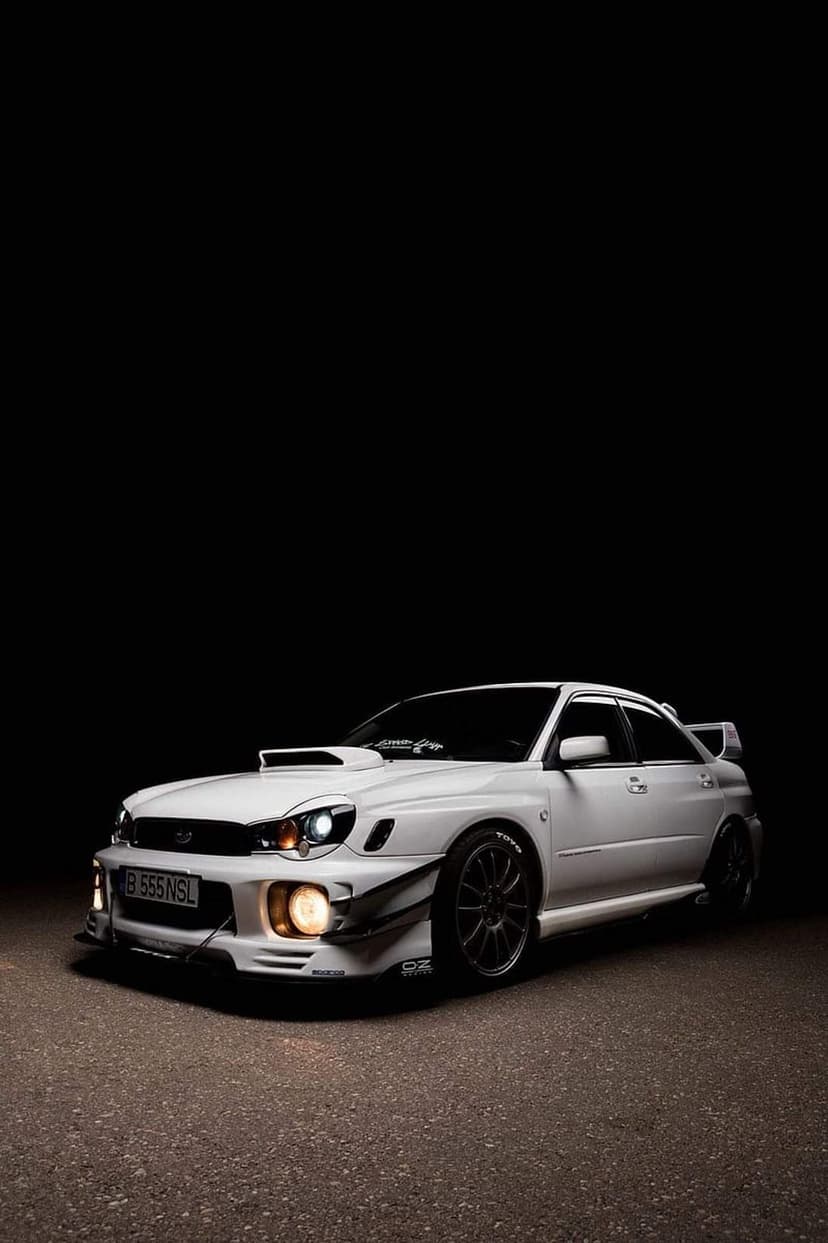 White Subaru WRX STI with glowing headlights and a large rear wing parked on dark asphalt under night skies.