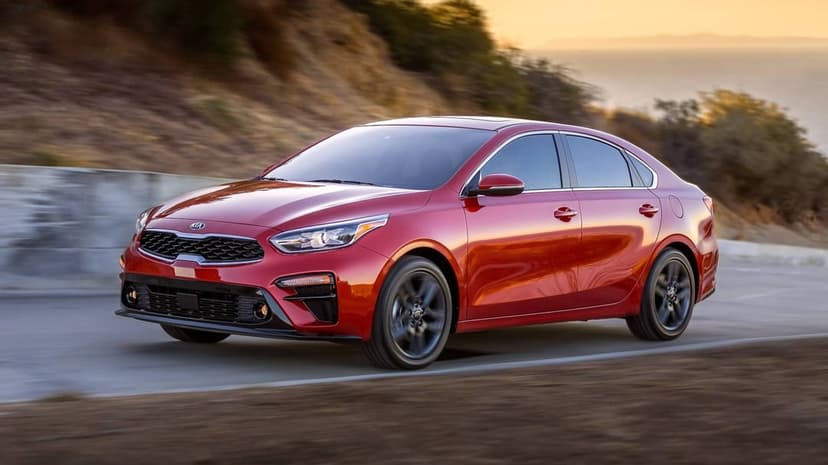 Red Kia Forte sedan driving on a winding coastal road during sunset with golden hour light reflections.