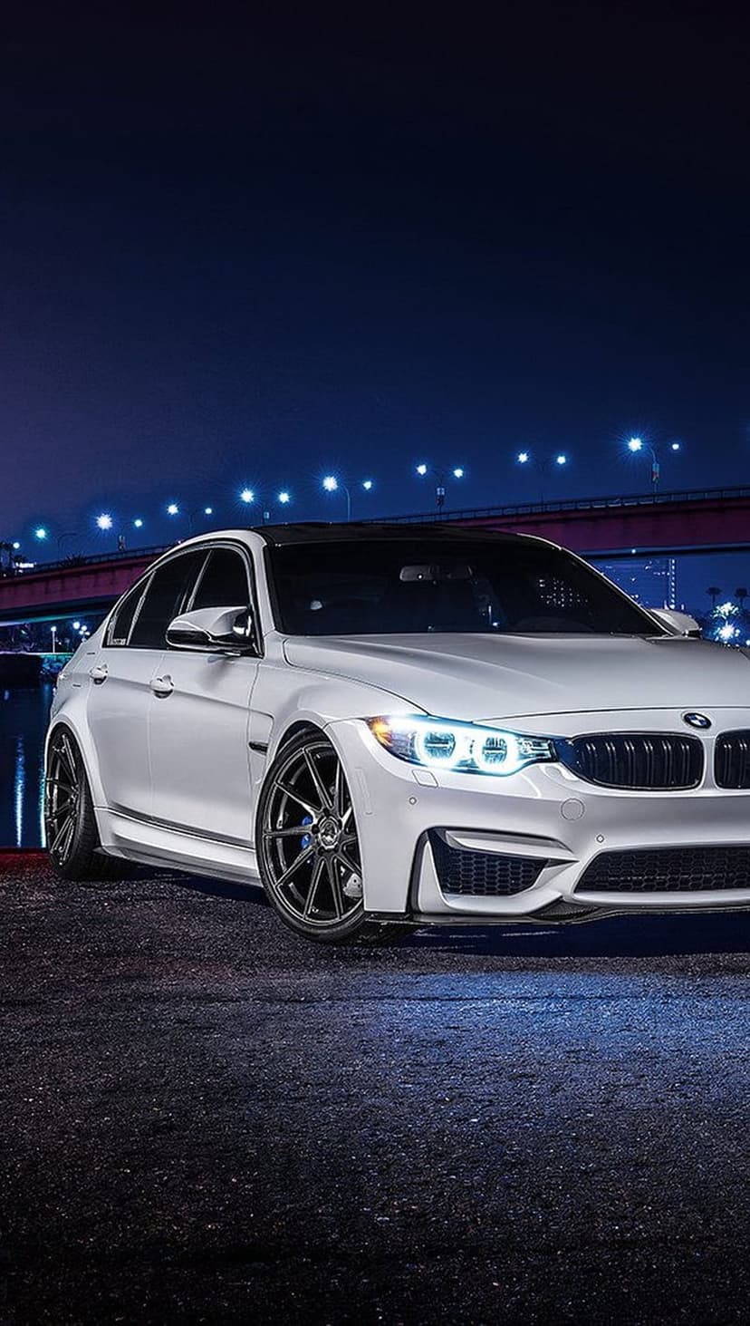 White BMW M3 sedan with glowing headlights parked on a city street at night near a bridge and shimmering water.
