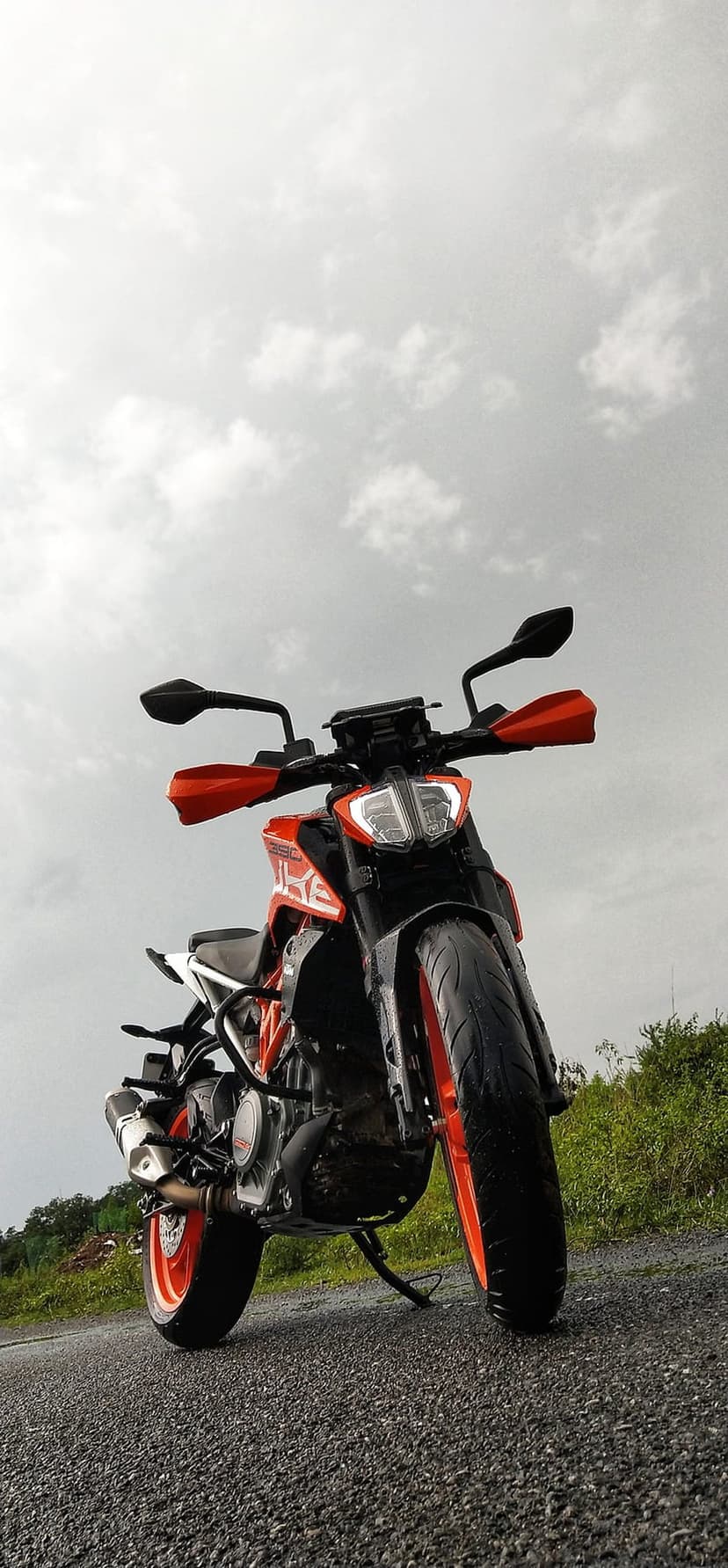 KTM Duke motorcycle with lit headlights stands on wet asphalt under a cloudy sky with green trees behind.