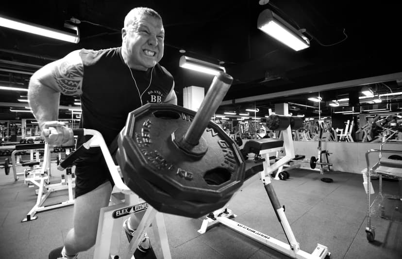 Muscular man with tattoos exerts force while lifting heavy circular weight plates in a dark, high-contrast gym.