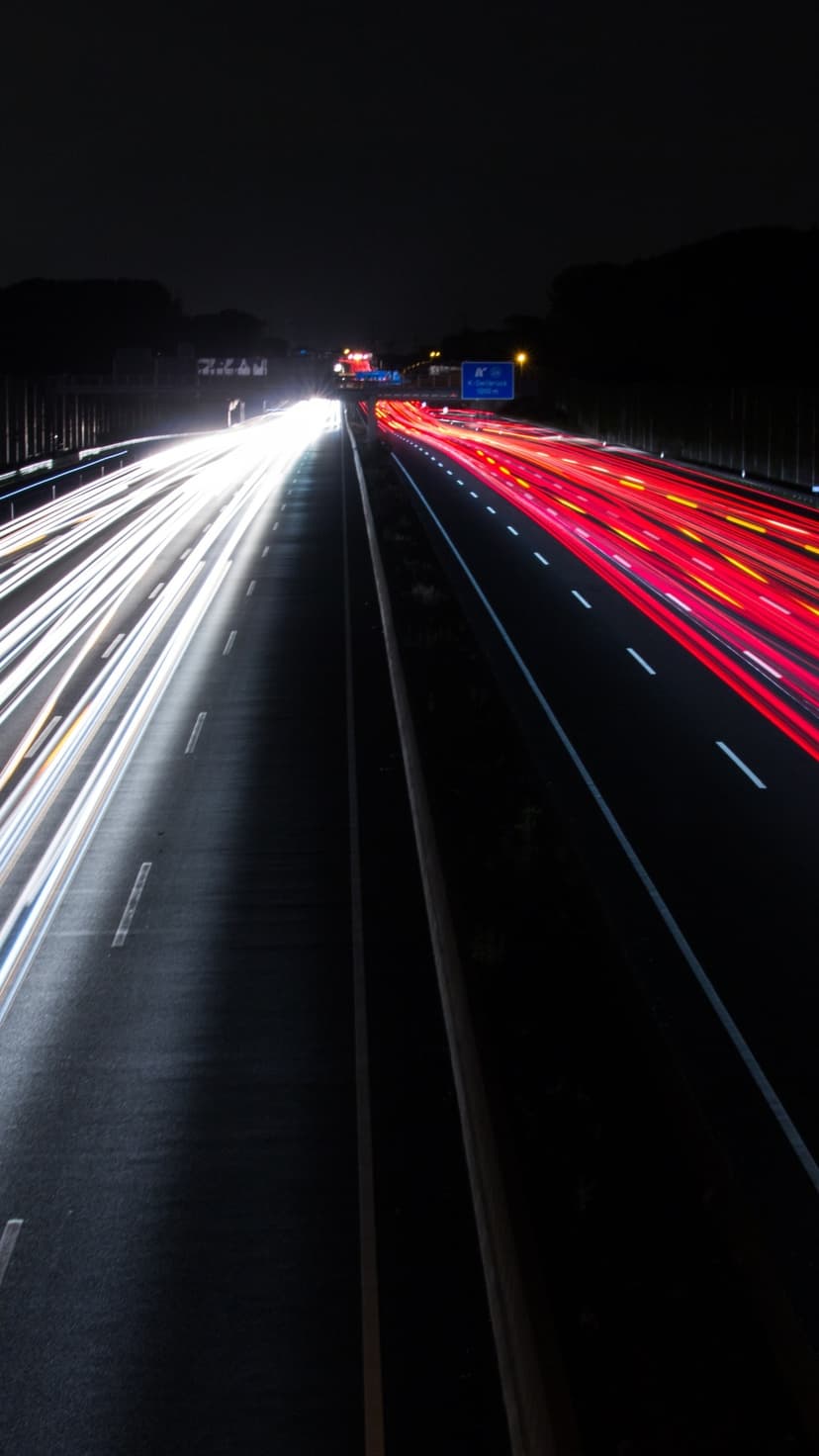 Red and white light trails stretch along a dark highway at night with a distant city glow in the background.