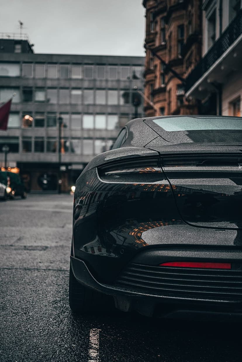 Rear view of a black sports car parked on wet asphalt with city lights reflecting on the glossy body at dusk.