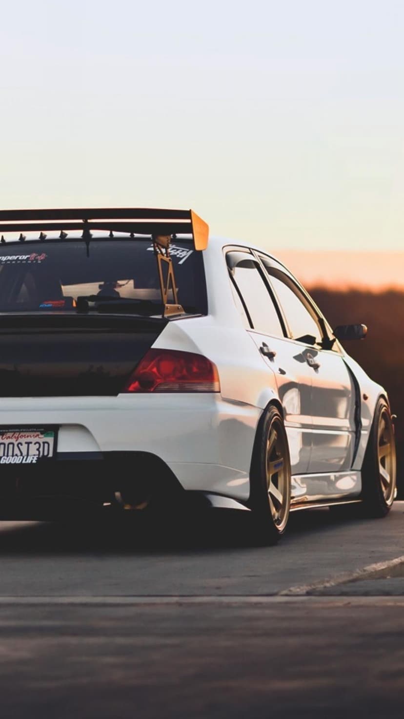 Rear view of a white Mitsubishi Lancer Evo with a large spoiler and gold rims during a golden sunset.