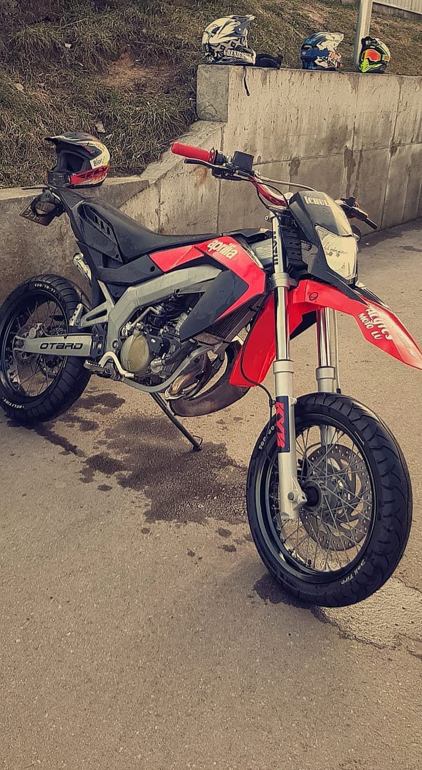 Red Aprilia supermoto motorcycle with red handlebars parked on wet concrete near motocross helmets in the background.