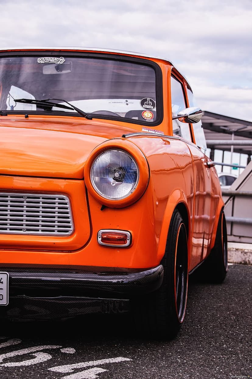 Orange Trabant 601 vintage car with custom wheels and front grille positioned against a cloudy sky backdrop.