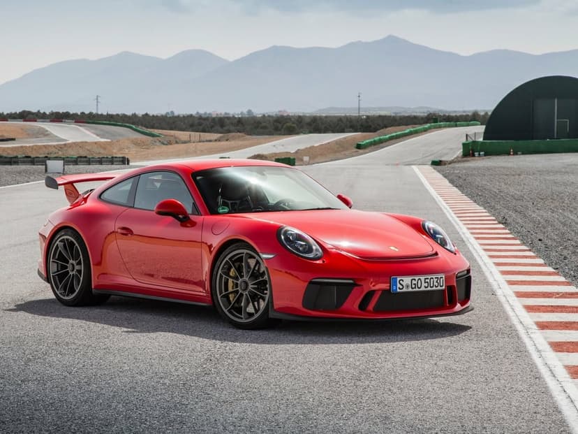Red Porsche 911 GT3 sports car on an asphalt racetrack with mountains and a hazy sky in the background.