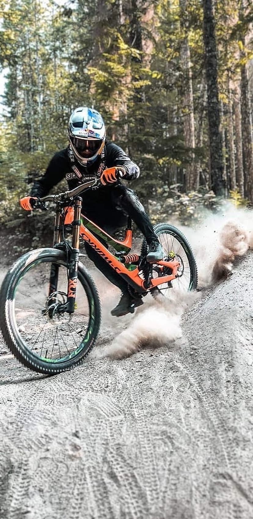 Mountain biker in orange gear leans into a dusty turn on a forest trail with tall trees in the background.