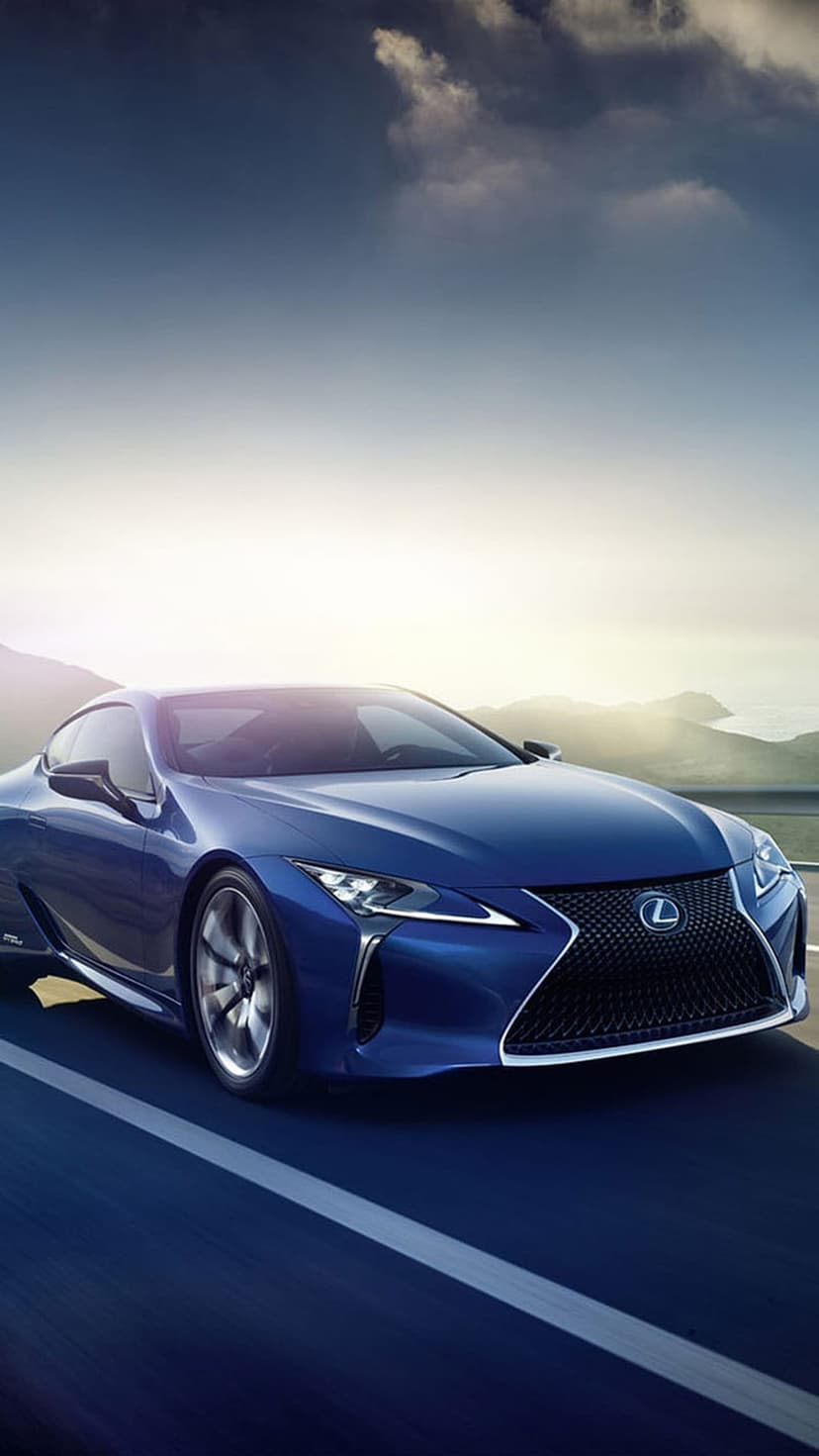 Blue Lexus LC coupe drives along a winding coastal road with hazy mountains and soft sunlight in the distance.