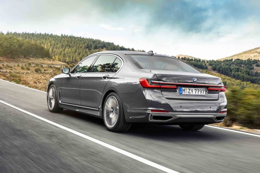 Grey BMW 750Li driving on a winding mountain road with green hills and a cloudy sky in the background