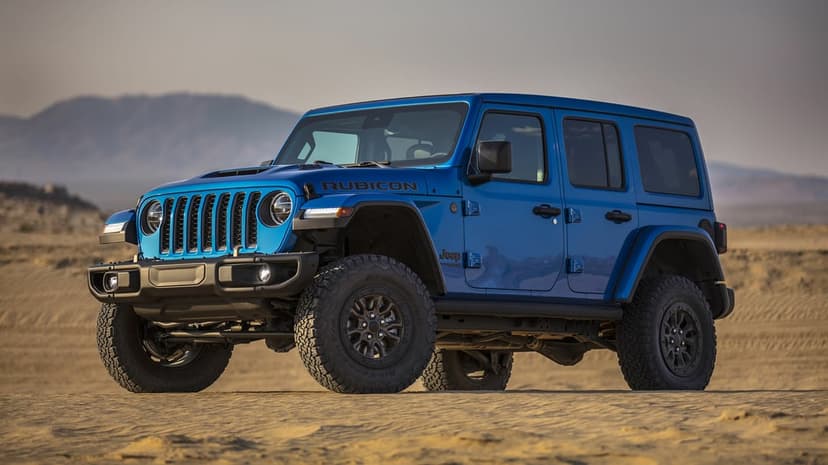 Blue Jeep Wrangler Rubicon parked on rippled sand dunes with mountains and a hazy sky in the background.