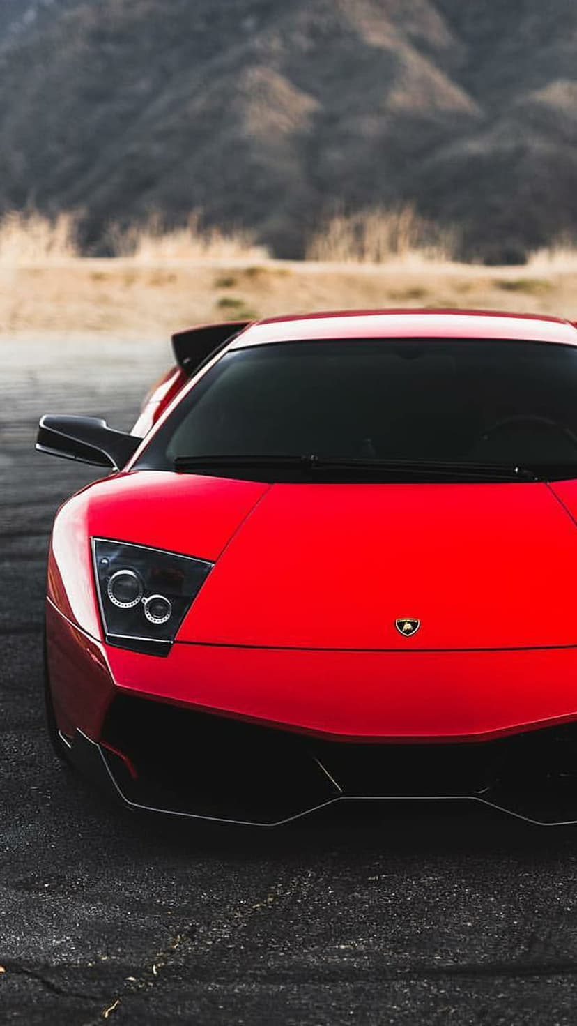 Red Lamborghini supercar with illuminated headlights parked on an asphalt road with blurred mountains behind it.