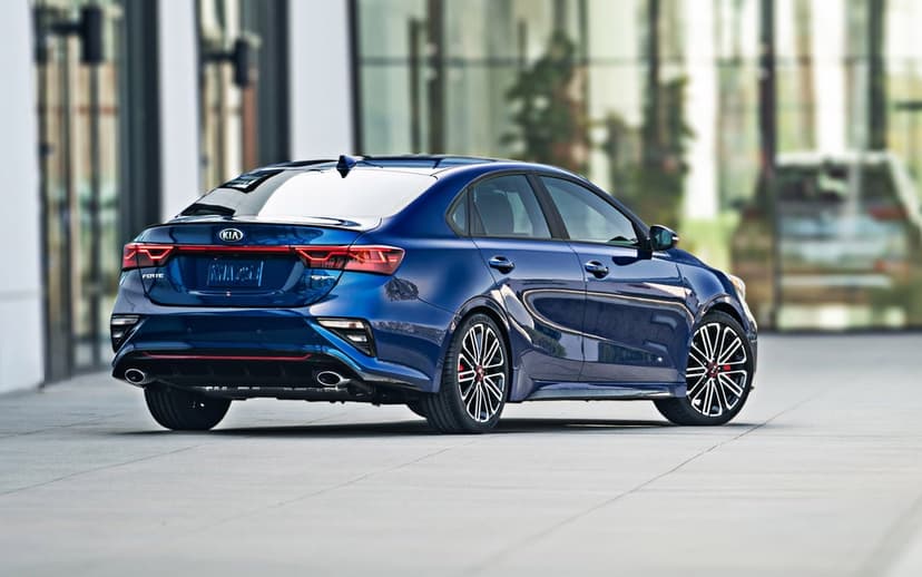 Blue Kia Forte GT sedan viewed from the rear on a grey plaza in front of a glass building with reflections.