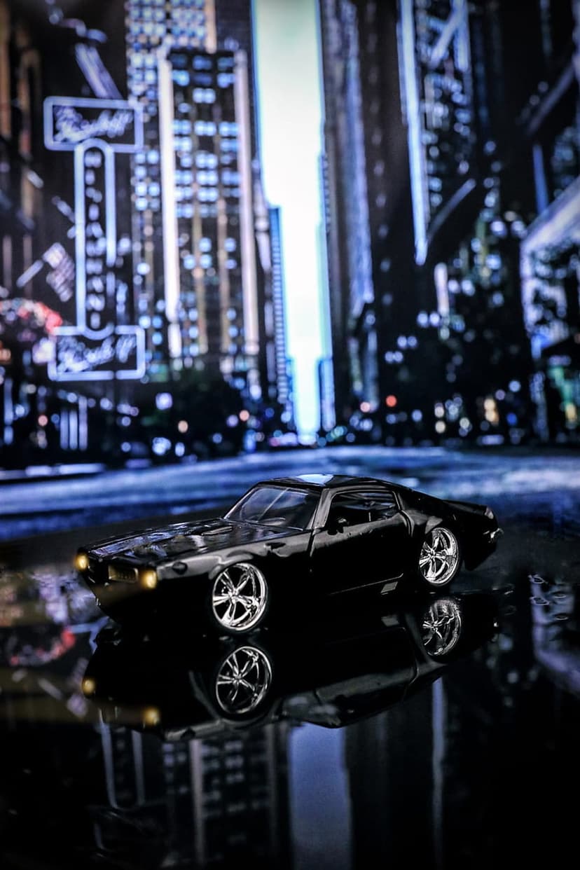 Black muscle car miniature on a wet night street with city light reflections and blurred urban skyscrapers.