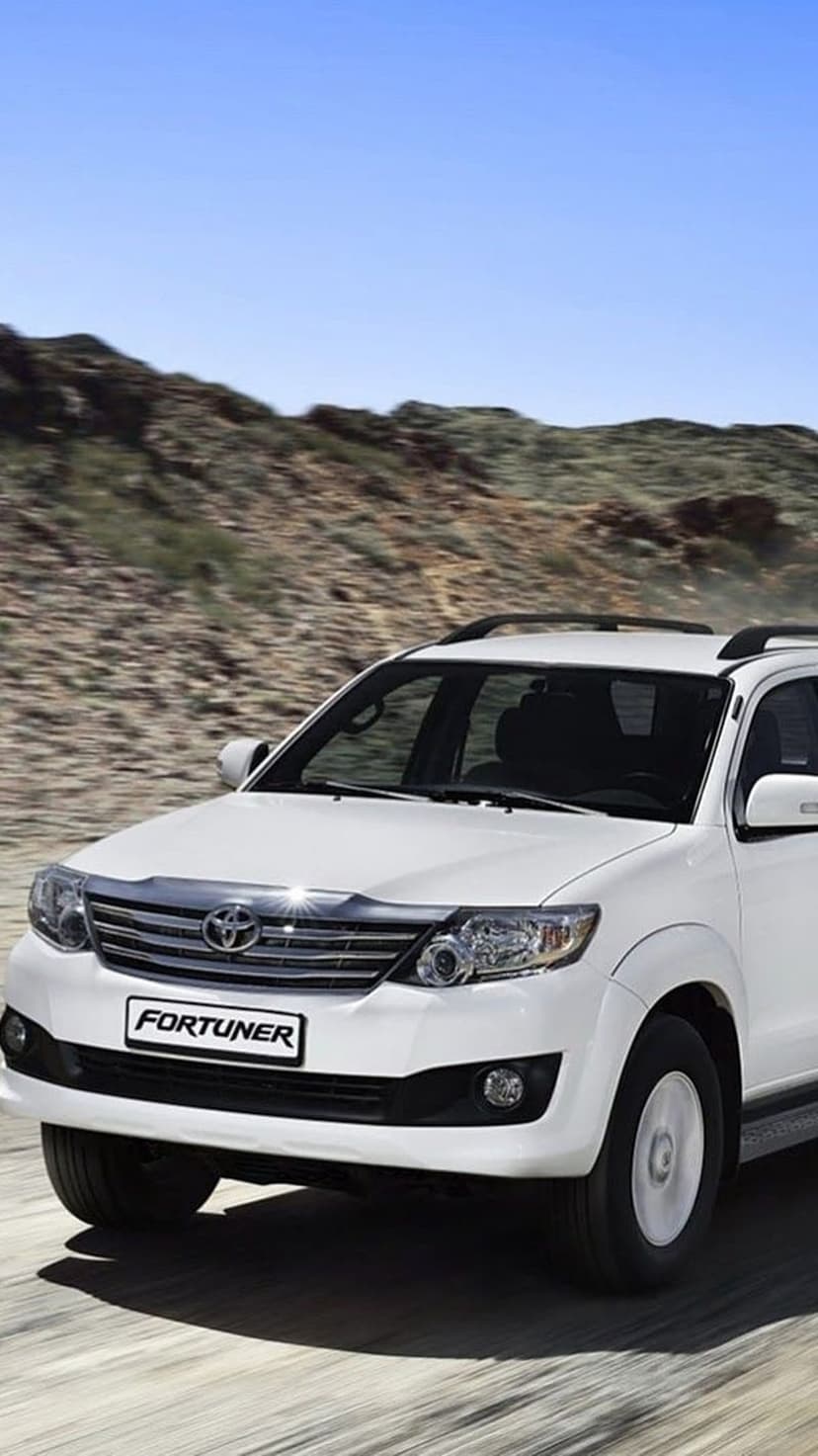 White Toyota Fortuner SUV kicks up a cloud of dust while driving through a rugged hilly landscape under a blue sky.