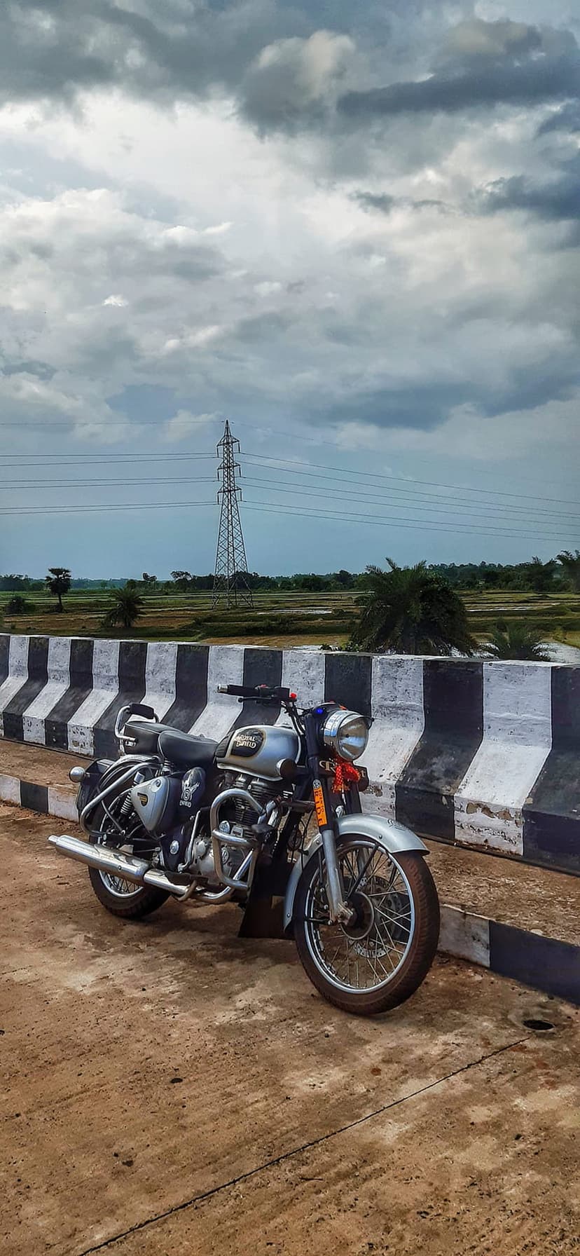 Silver Royal Enfield motorcycle on a bridge with striped barriers overlooking rural fields under a cloudy sky.