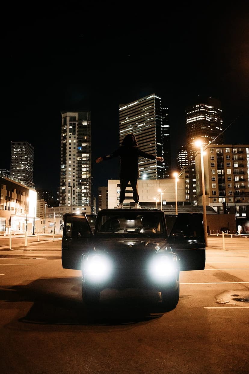 Person in a hoodie standing on a car roof with arms wide open against a backdrop of illuminated city skyscrapers.
