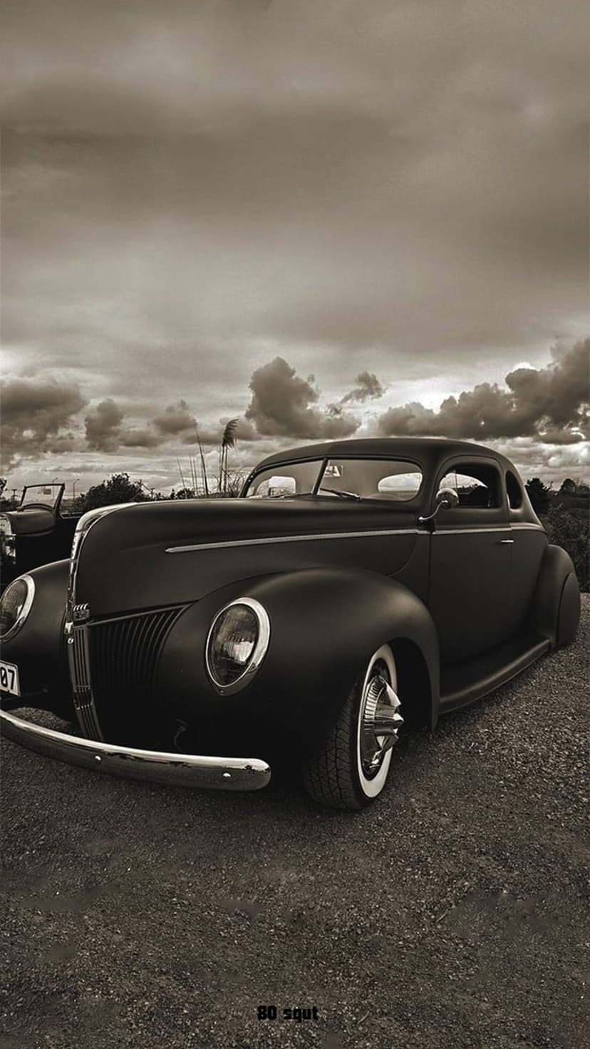 1940 Ford Coupe in black and white parked on a gravel road beneath a dramatic sky filled with dark storm clouds.