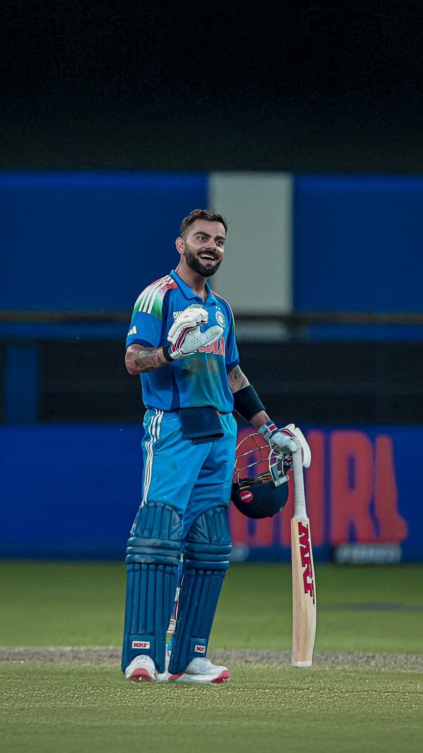 Virat Kohli smiles on a cricket pitch wearing a blue jersey, holding a bat and helmet against a blurred stadium.