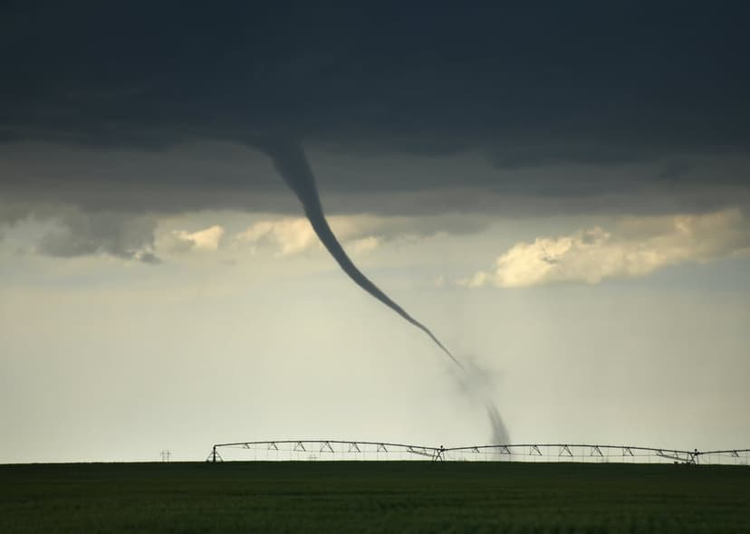 Towering tornado funnel touching down on a green field next to a long metal irrigation pipe under dark clouds.